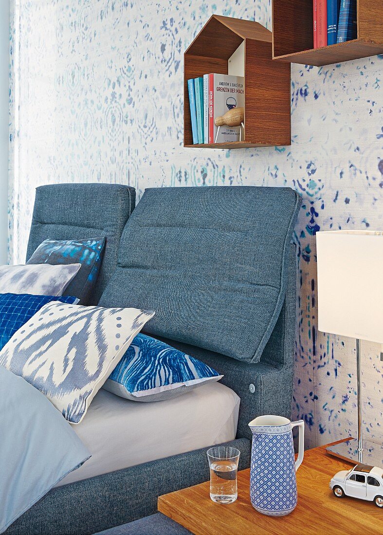 Doppelbett und Bettkopfteil mit Denimbezug vor Tapete mit verschwommenen Mustern, darüber kleine Regalmodule in Häuschenform