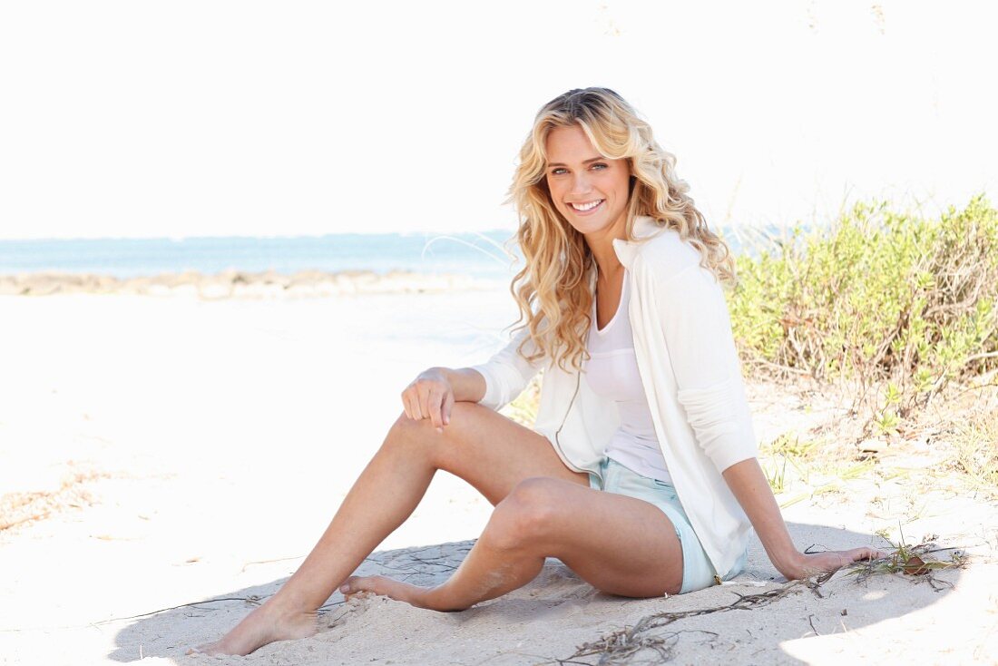 A young blonde woman on a beach wearing a white top, jacket and shorts