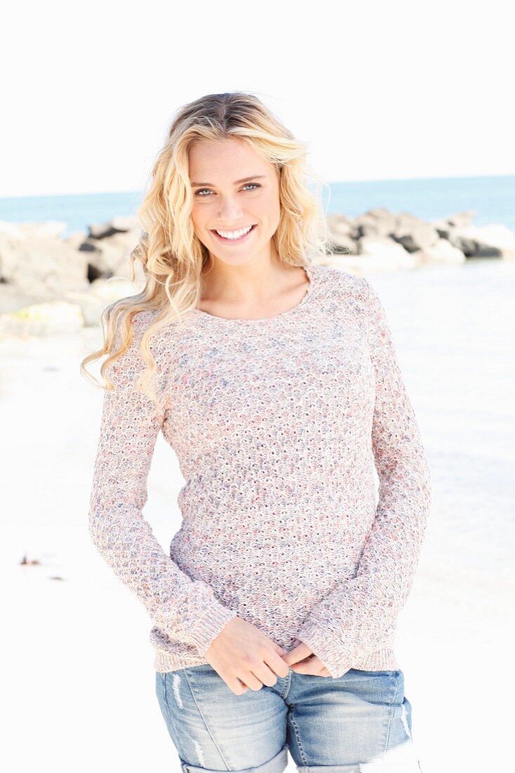 A young blonde woman wearing a knitted jumper and jeans by the sea