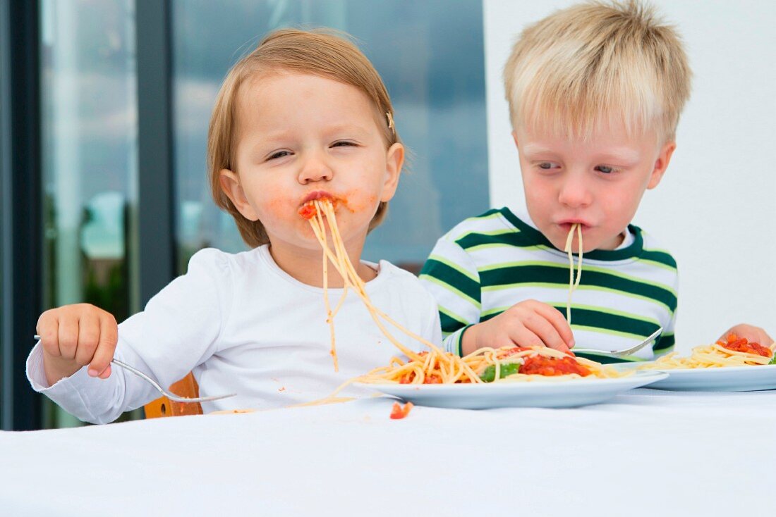 Two small children eating spaghetti with tomato sauce
