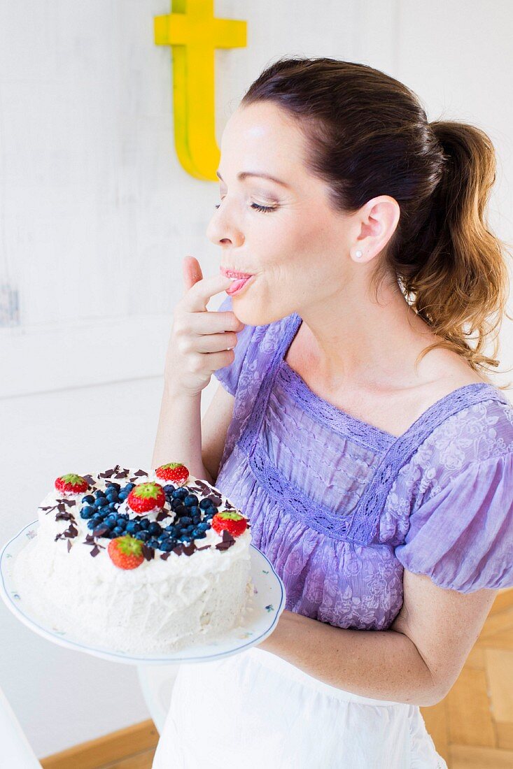 A woman holding a berry cream cake licking cream from her finger