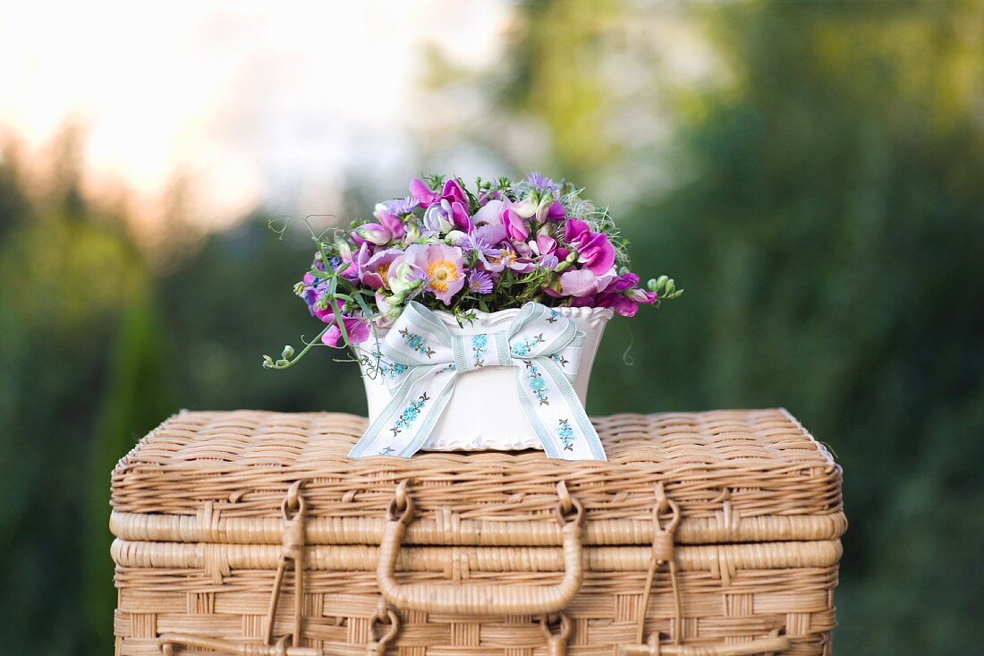 Pinkfarbenes Blumenarrangement in Porzellanschale auf Weidenkoffer