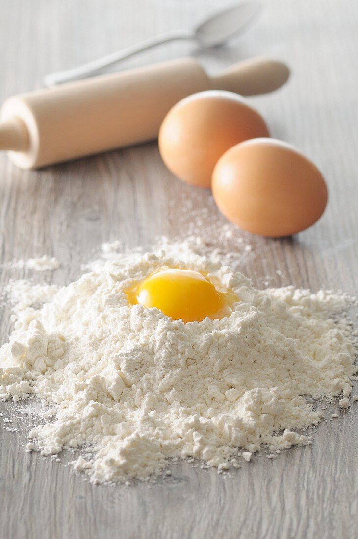 An egg yolk in a pile of flour with whole eggs and a rolling pin