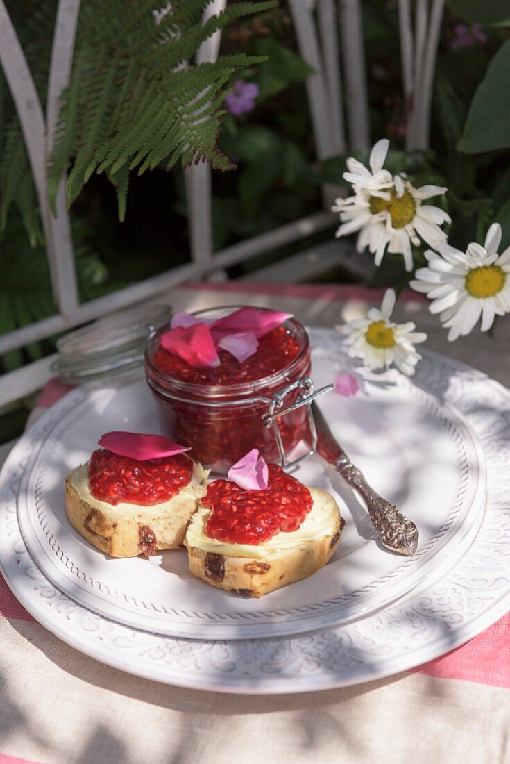 Rosinenbrötchen mit Himbeermarmelade und Rosenblütenblättern auf Gartentisch