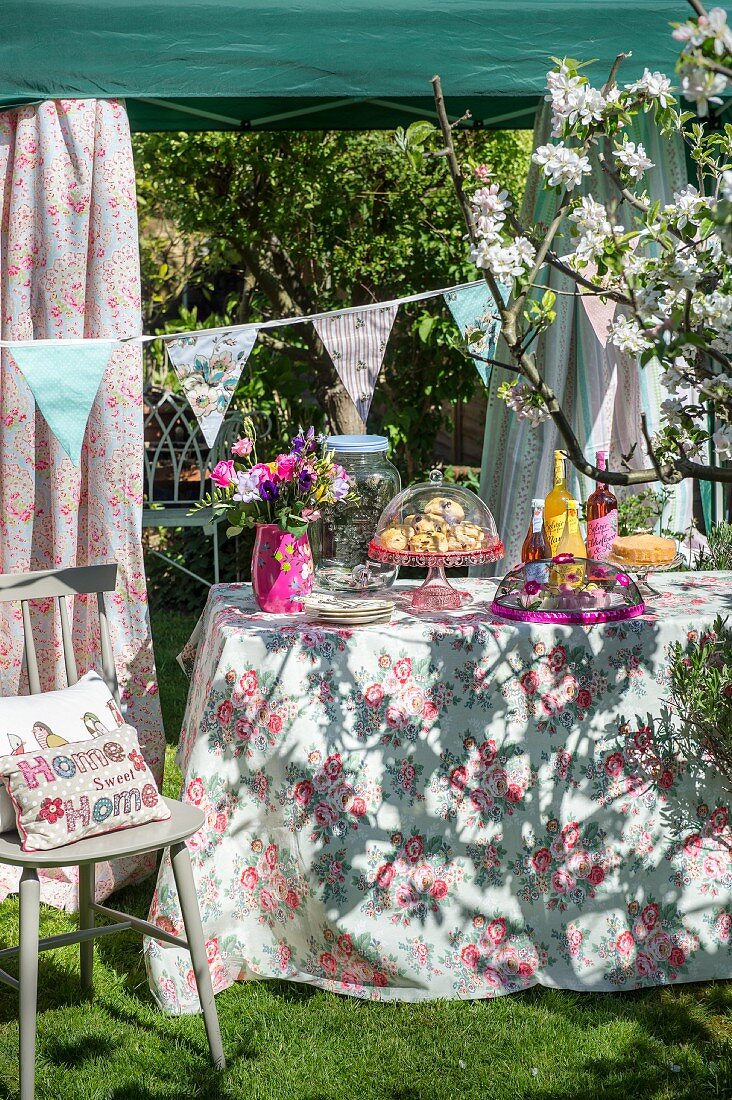 Garden buffet in sunshine on floral tablecloth below pavilion decorated with bunting
