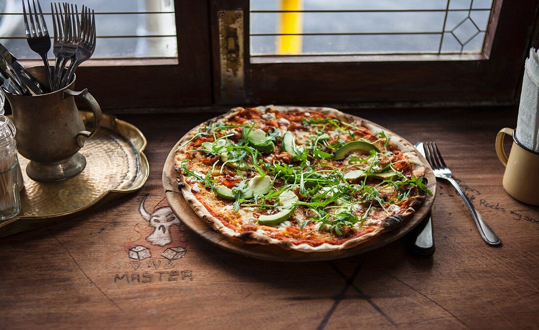 Pizza mit Avocado und Rucola auf Holztisch vor Fenster in Restaurant