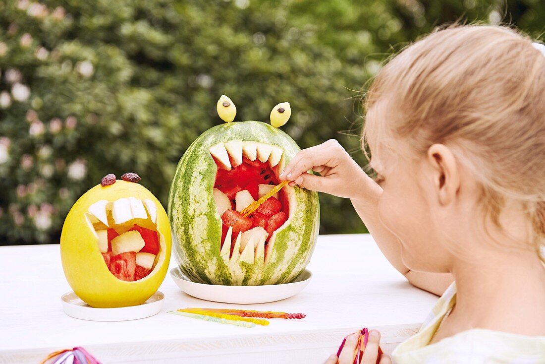 Melonenwürfel in ausgehöhlten Melonenschalen, Mädchen nimmt sich ein Stück mit Spiesschen