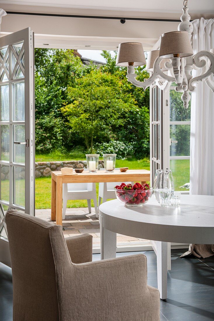 Sessel mit heller Husse an weißem Tisch unter Kronleuchter, vor offenen Terrassentüren mit Blick auf Terrasse