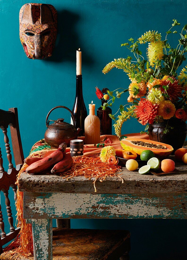 A bunch of flowers and fruit on a wooden table with a wooden mask in the background as wall decoration