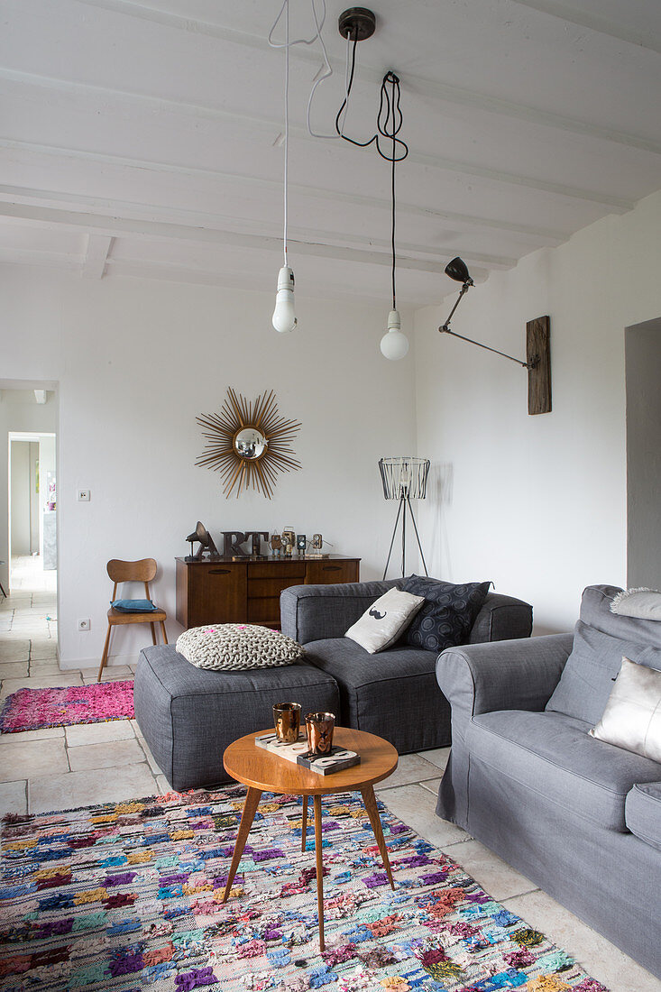 Retro side table and seating with grey upholstery in simple living room with bare bulbs hanging from white wood-beamed ceiling