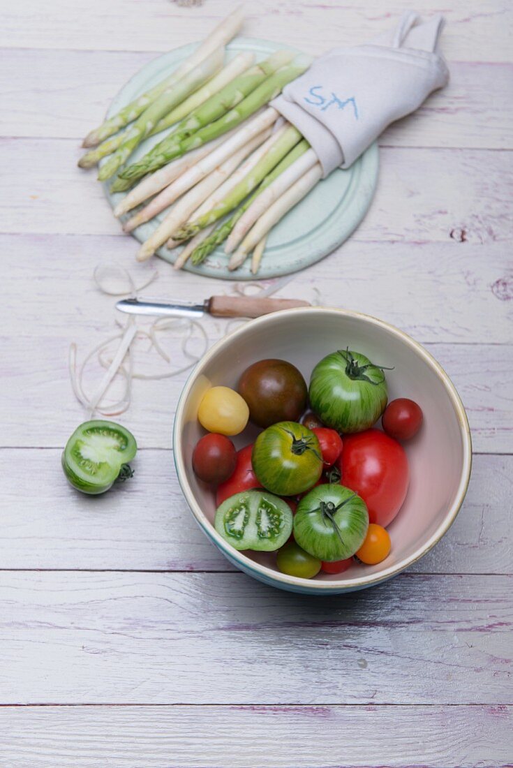 Weisser und grüner Spargel auf Holzbrett, Spargelschalen, Schäler und Schüssel mit verschiedenen Tomaten