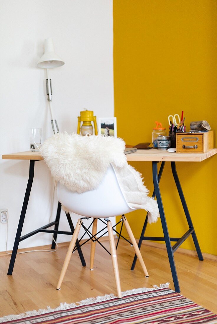 Desk with wooden top on metal frame and sheepskin on shell chair in corner against yellow wall