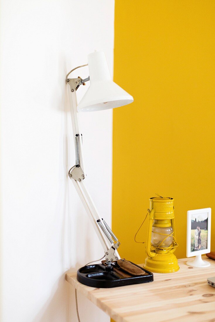 White desk lamp next to yellow storm lamp on wooden table in corner against yellow and white wall