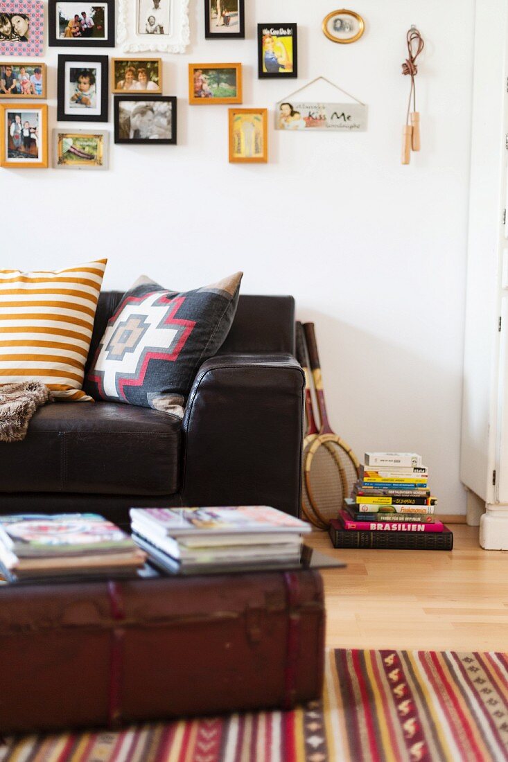 Stacked magazines on top of vintage trunk in front of leather sofa below gallery of family photos