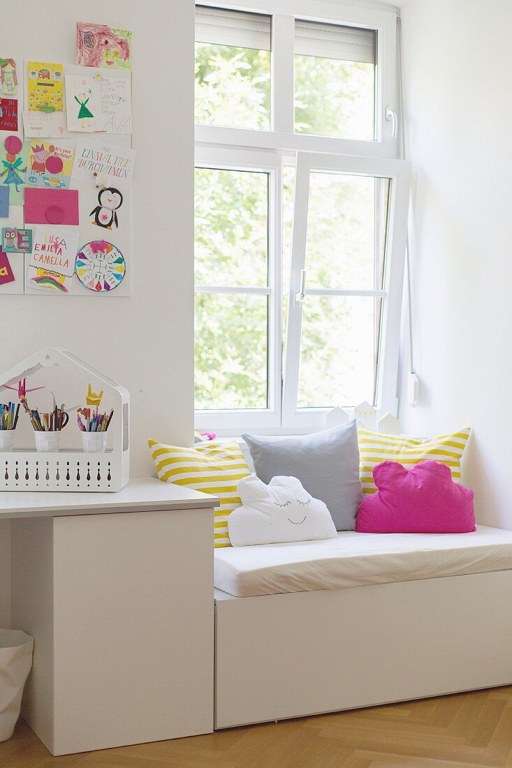 Weißer Schreibtisch vor Wand mit bunten Kinderzeichnungen, eingebaute Fenstersitzbank mit Sitzpolster und Kissen