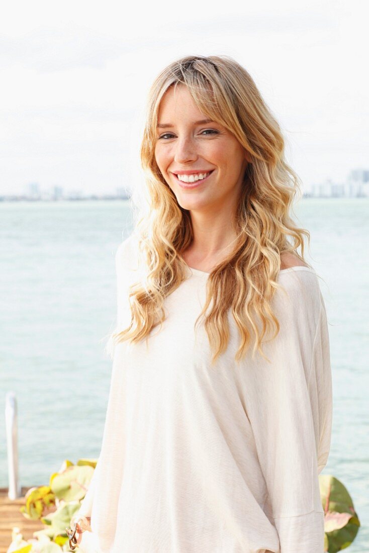 A young blonde woman wearing a light, oversized shirt by the sea