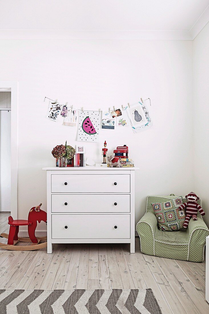 Kinderzimmer mit weisser Schubladenkommode, Schaukeltier und grünem Kindersessel, Wanddekoration mit Zeichnungen und Fotos an Wäscheleine