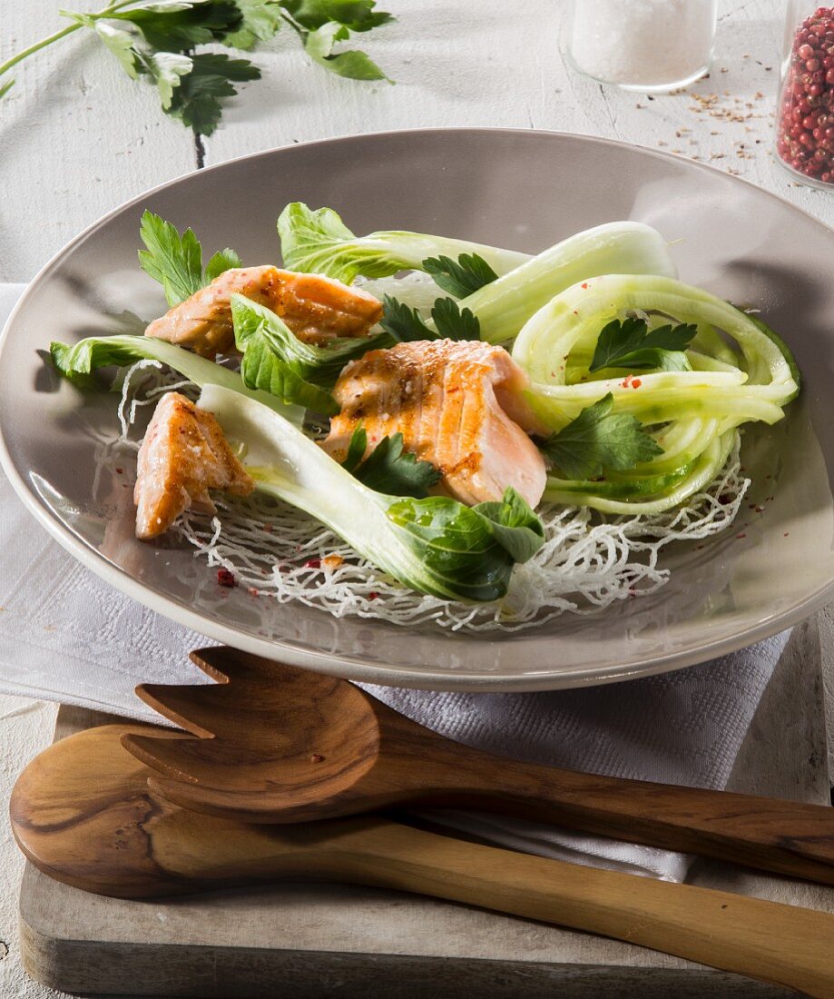 Salmon and bok choy on a bed of glass noodles on a plate with wooden cutlery
