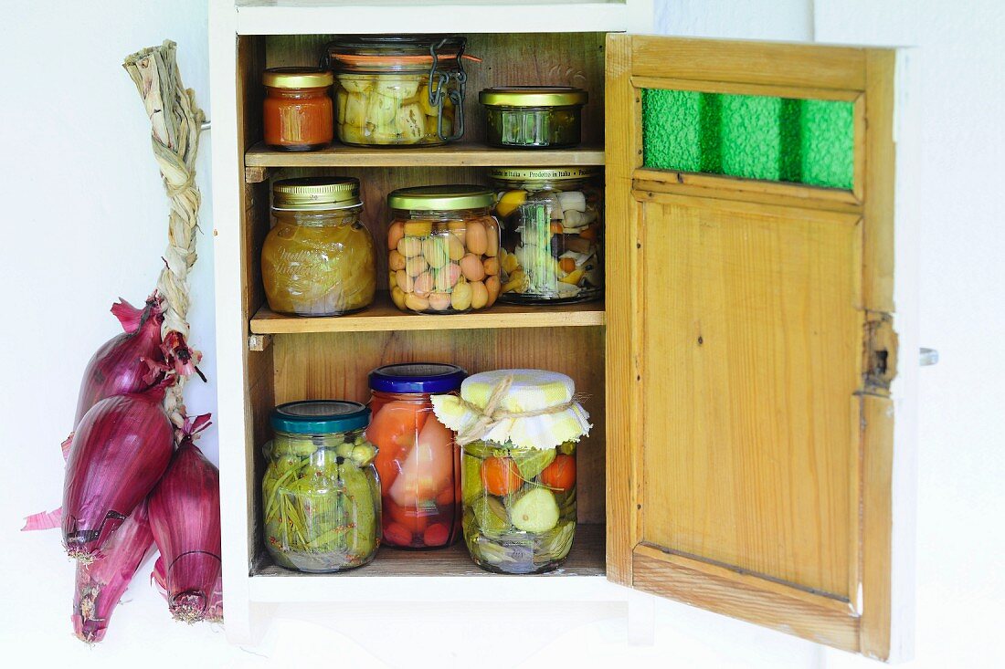 Eingmachtes Gemüse in Gläsern im Wandschränkchen mit offener Tür, daneben aufgehängte Zwiebeln
