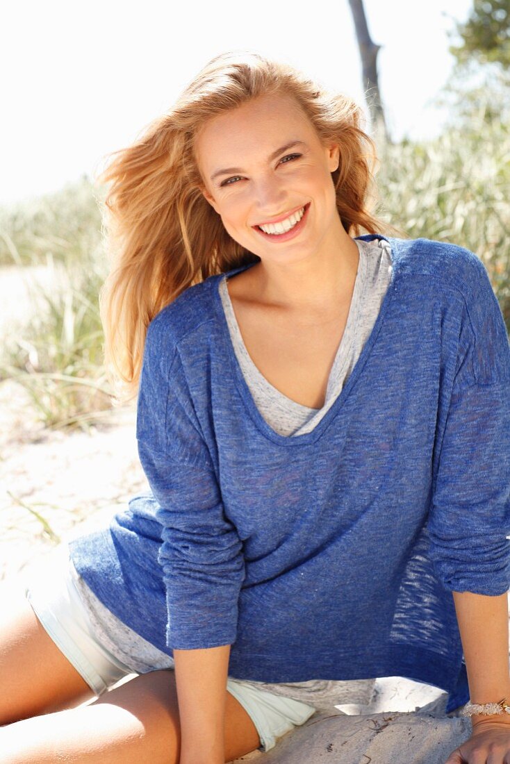 A young woman wearing shorts and a blue mottled shirt sitting on a beach