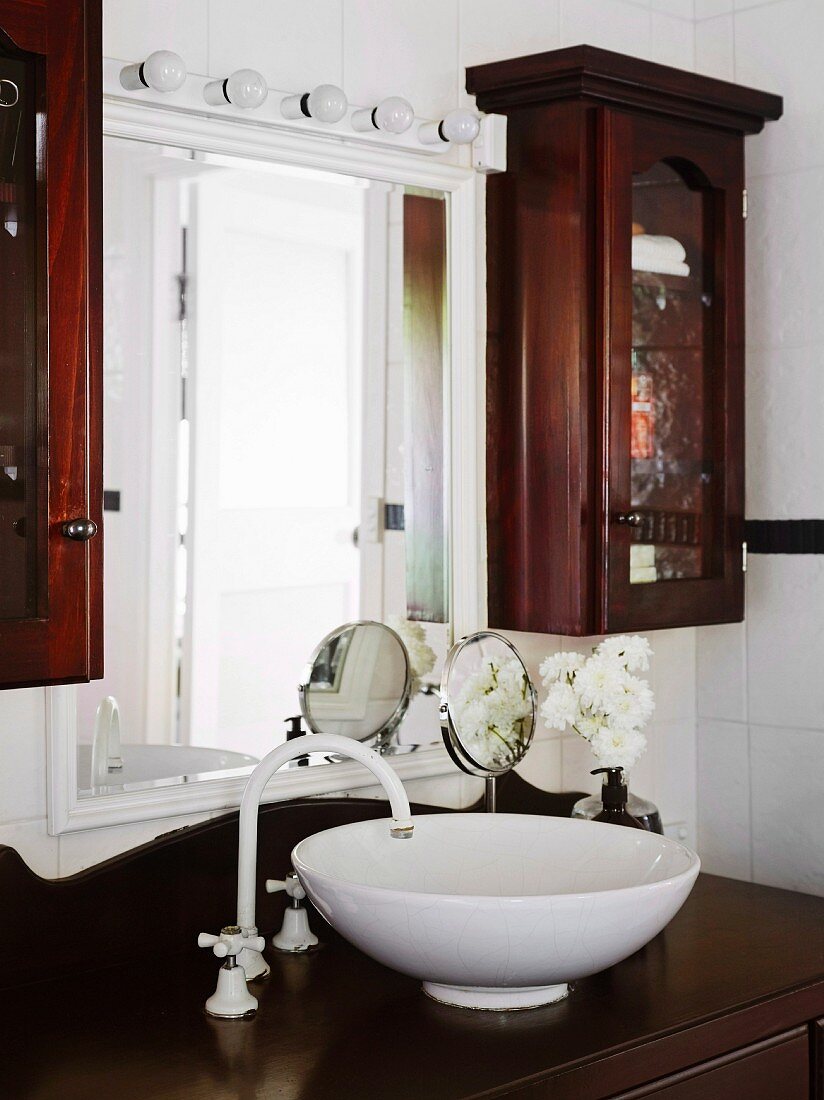 White washbowl on dark brown wooden base cabinet in front of wall mirror and narrow display cabinets