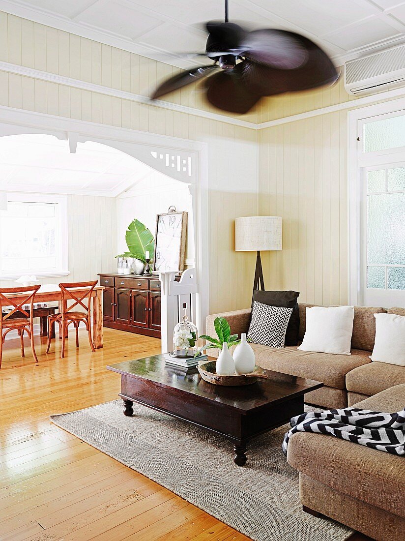 Dark brown wood coffee table and corner sofa in wood-clad living room with wide passage and view of dining area