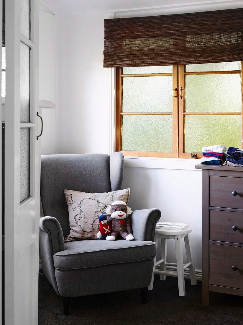 View through open room door to gray reading armchair with soft toys and pillows, next to lattice window with bamboo blind in the children's room