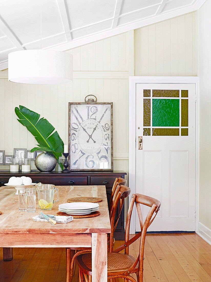 Wooden dining table and wooden chairs with wicker in front of a framed vintage clock on a sideboard in the attic