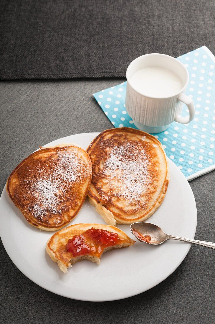 Buttermilch-Pancakes mit Puderzucker und Erdbeermarmelade auf Teller vor einer Tasse Milch