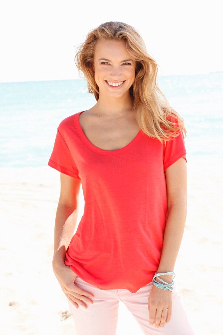 A blonde woman on a beach wearing a red top and pink trousers