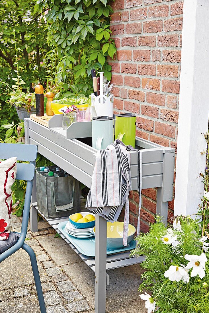 Grau lackierter Pflanztisch als Anrichte mit Thermoskannen, Kochutensilien und Geschirr auf der Terrasse