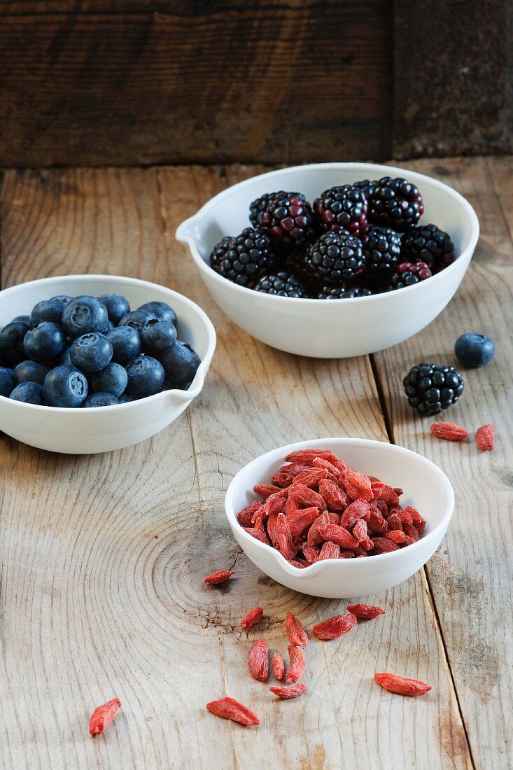 Goji berries, blackberries and blueberries in bowls on a wooden surface
