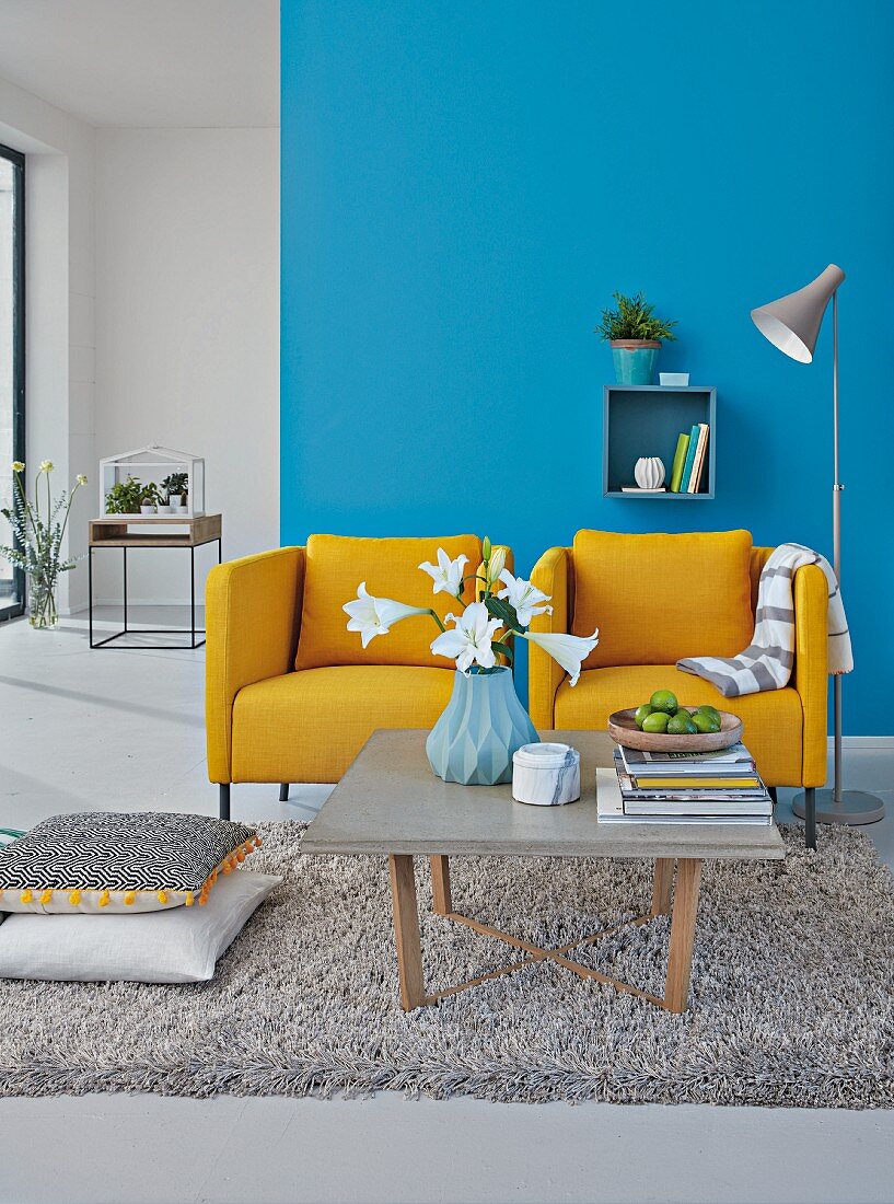 Two mustard-yellow upholstered armchairs against a blue wall with a coffee table with a concrete surface on a shag pile rug