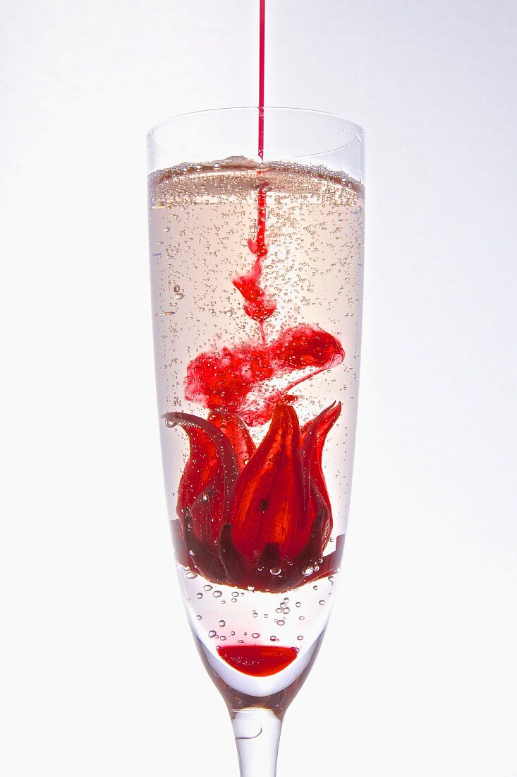 Red syrup being added to a hibiscus flower in a glass of champagne