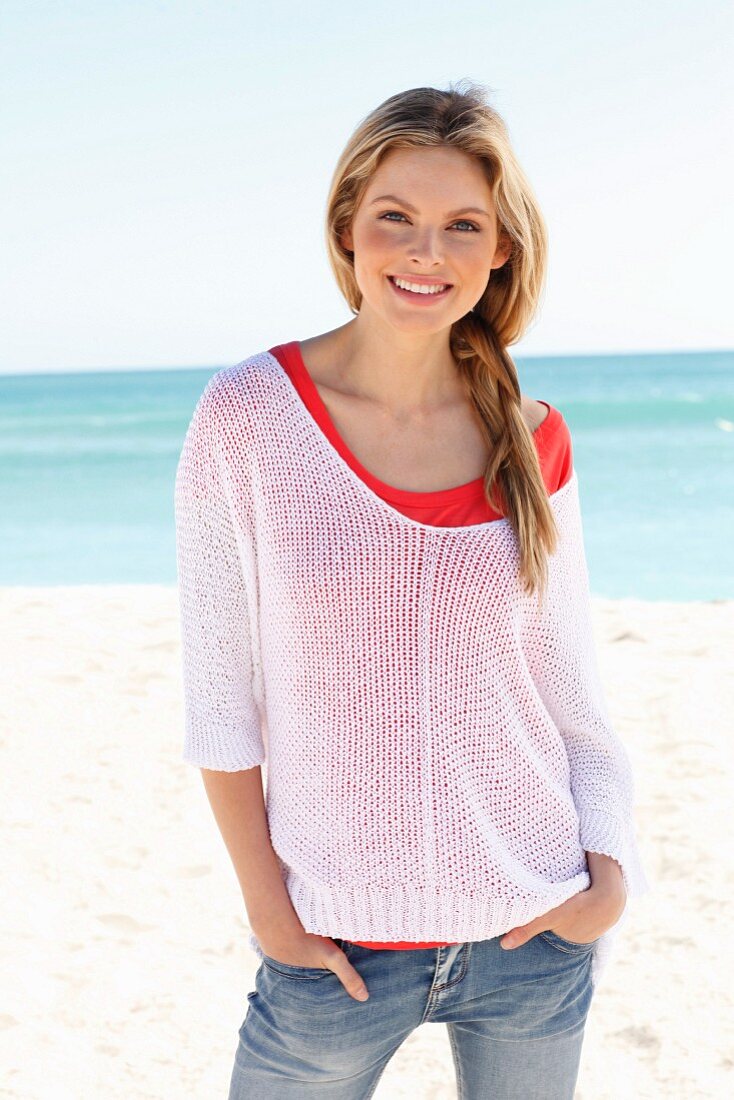 Junge Frau in rotem Shirt, weißem Lochstrickpulli und Jeans am Strand