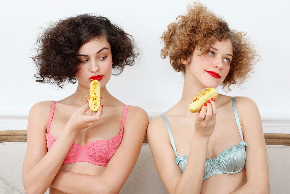 Two young women wearing bras and eating eclairs