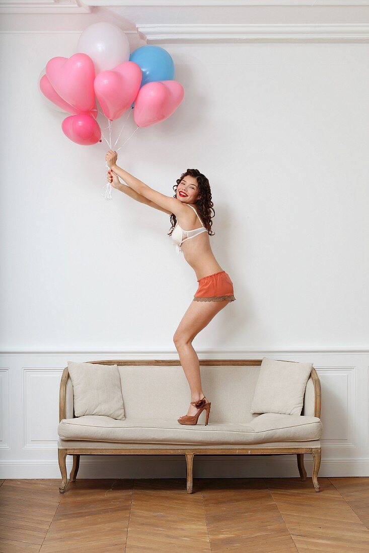 Junge Frau in Unterwäsche mit Luftballons steht auf Sofa