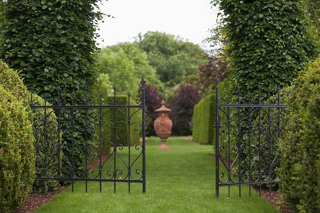 Wrought iron gate between tall, clipped box hedges and well-tended lawn path leading to amphora