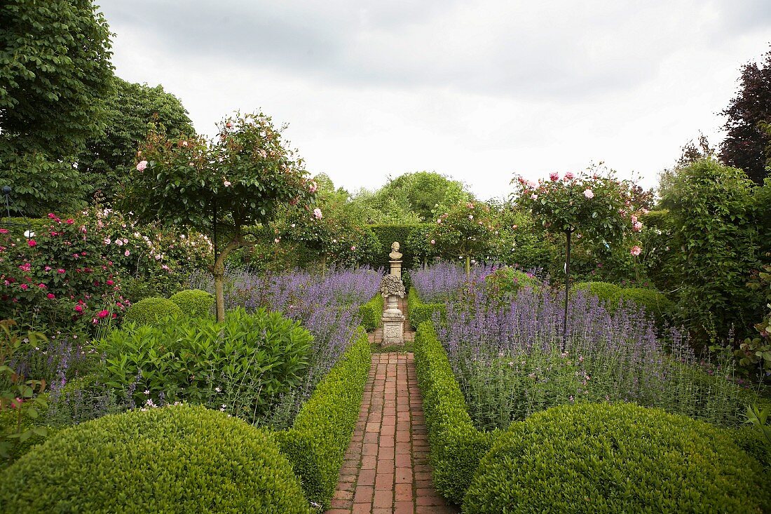 Brick-paved path leading through beds of clipped hedges, standard roses and box balls to stone sculptures on plinths