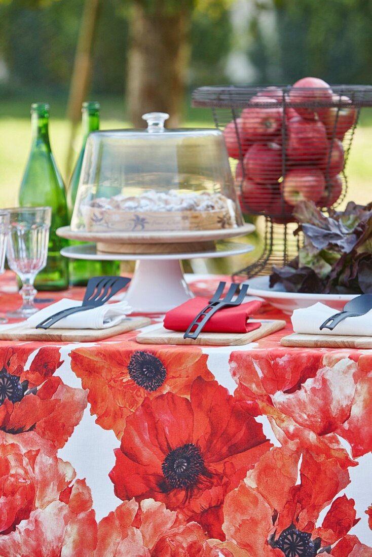 Red floral tablecloth, cake and basket of apples on table in garden