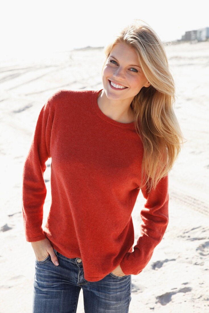 A young woman on a beach wearing a rust-red jumper and jeans