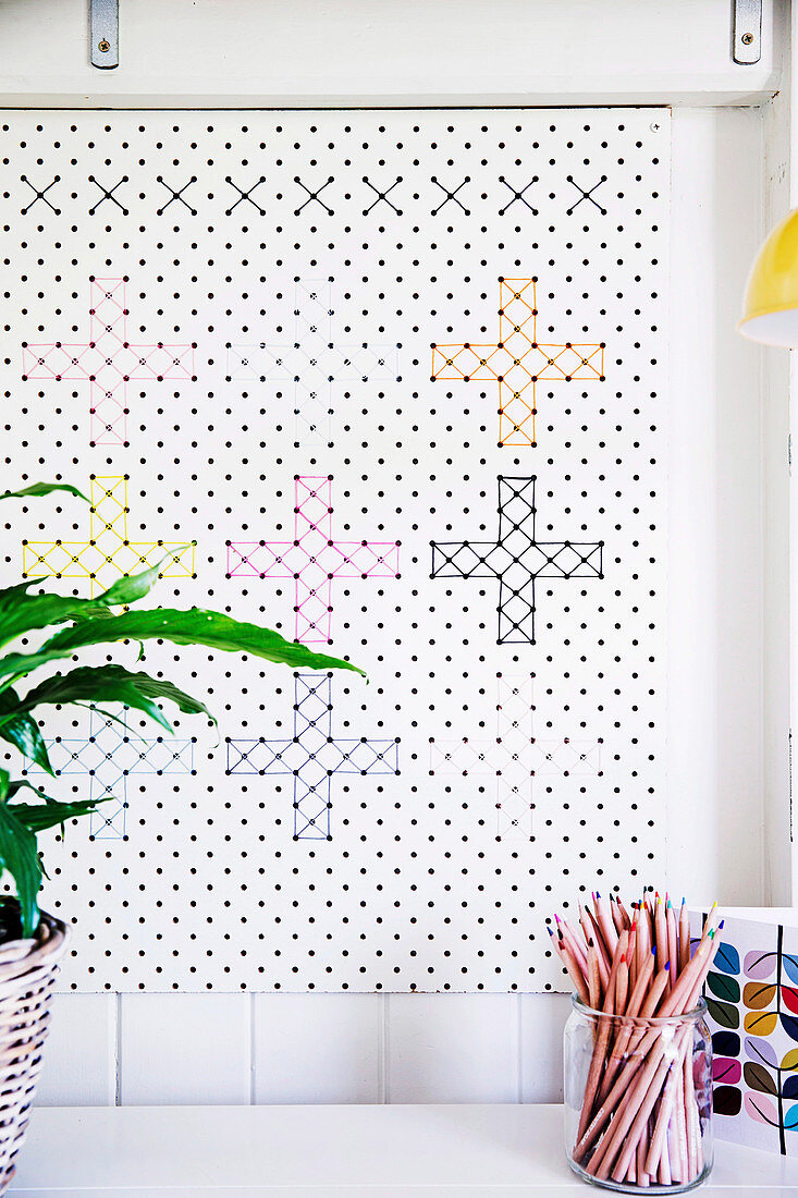 White perforated plate with cross-stitches in different colors as wall decoration, in front of glass with colored pencils