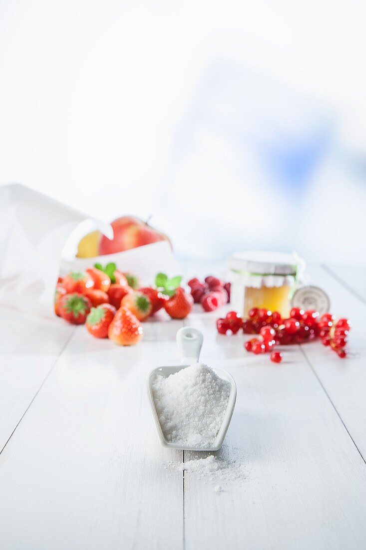 Schippe mit Zucker auf weißem Holz vor Früchtearrangement bestehend aus Erdbeeren, Preiselbeeren und Äpfeln