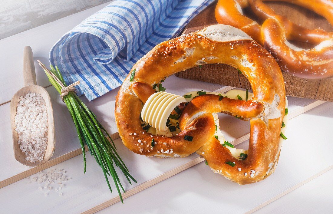 Laugenbrezel mit Butter und Schnittlauch neben Salzschaufel und Schnittlauchbündel auf weißem Holz