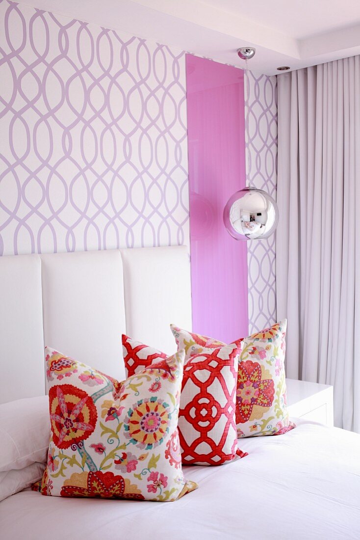 Three colourful scatter cushions on bed against wall covered in purple-patterned wallpaper
