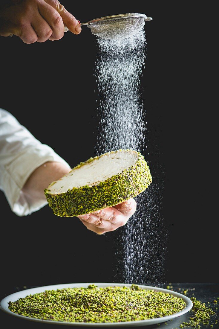 A confectioner dusting a small pistachio cake with icing sugar
