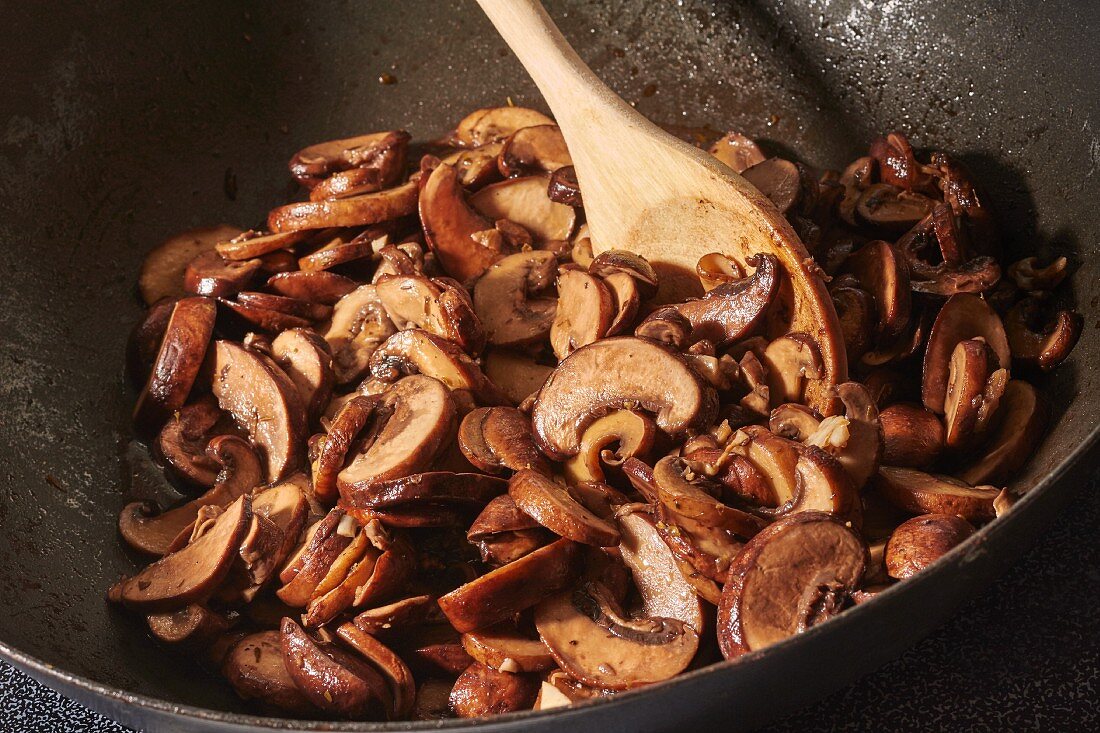 Brown mushrooms being fried with chopped garlic in olive oil in a wok