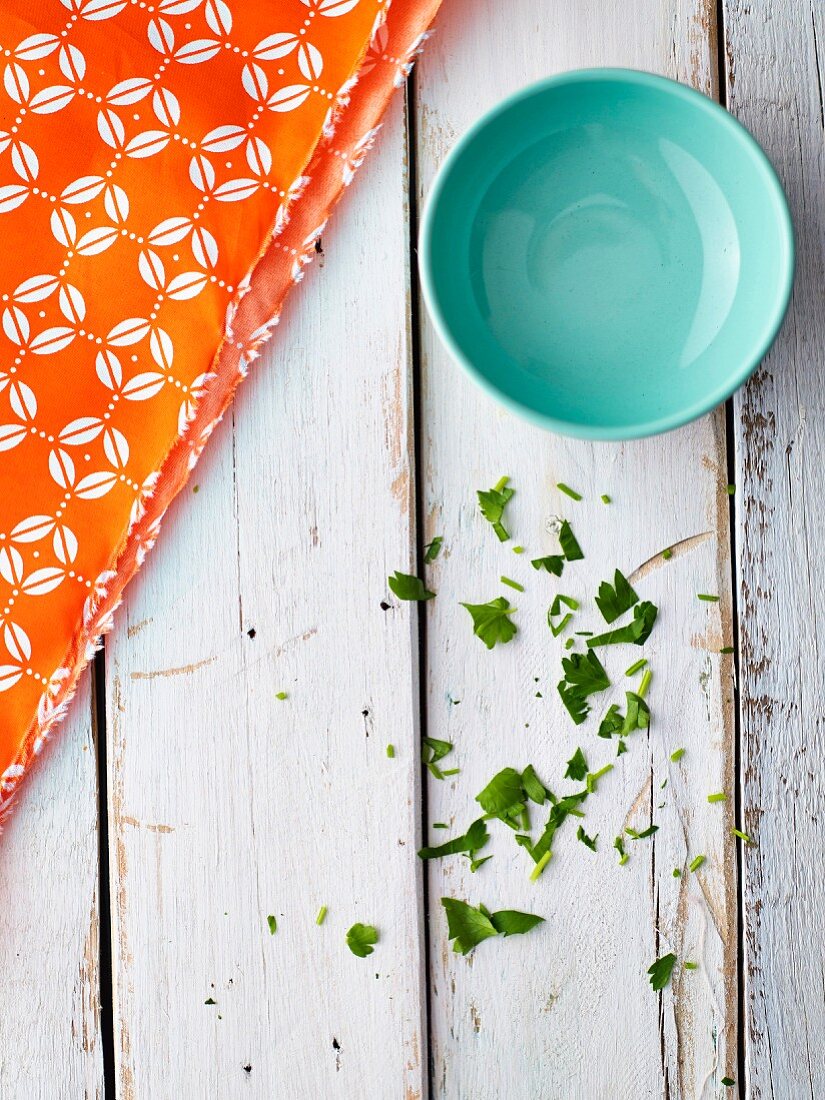 A blue bowl on a white shabby wooden table with a bright orange napkin and chopped parsley