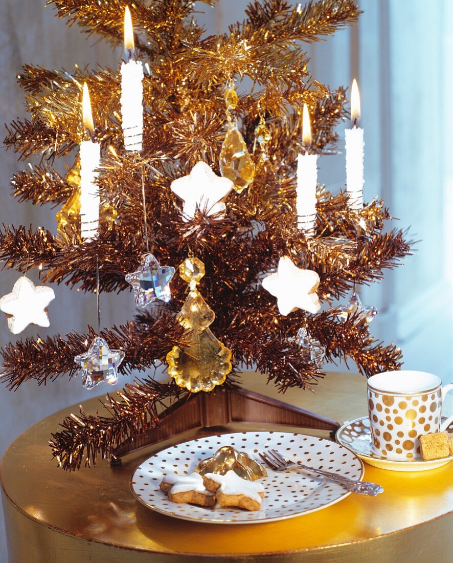 Goldfarbenes Deko-Weihnachtsbäumchen mit brennenden Kerzen auf glänzendem Beistelltisch und elegantem Kaffeegedeck