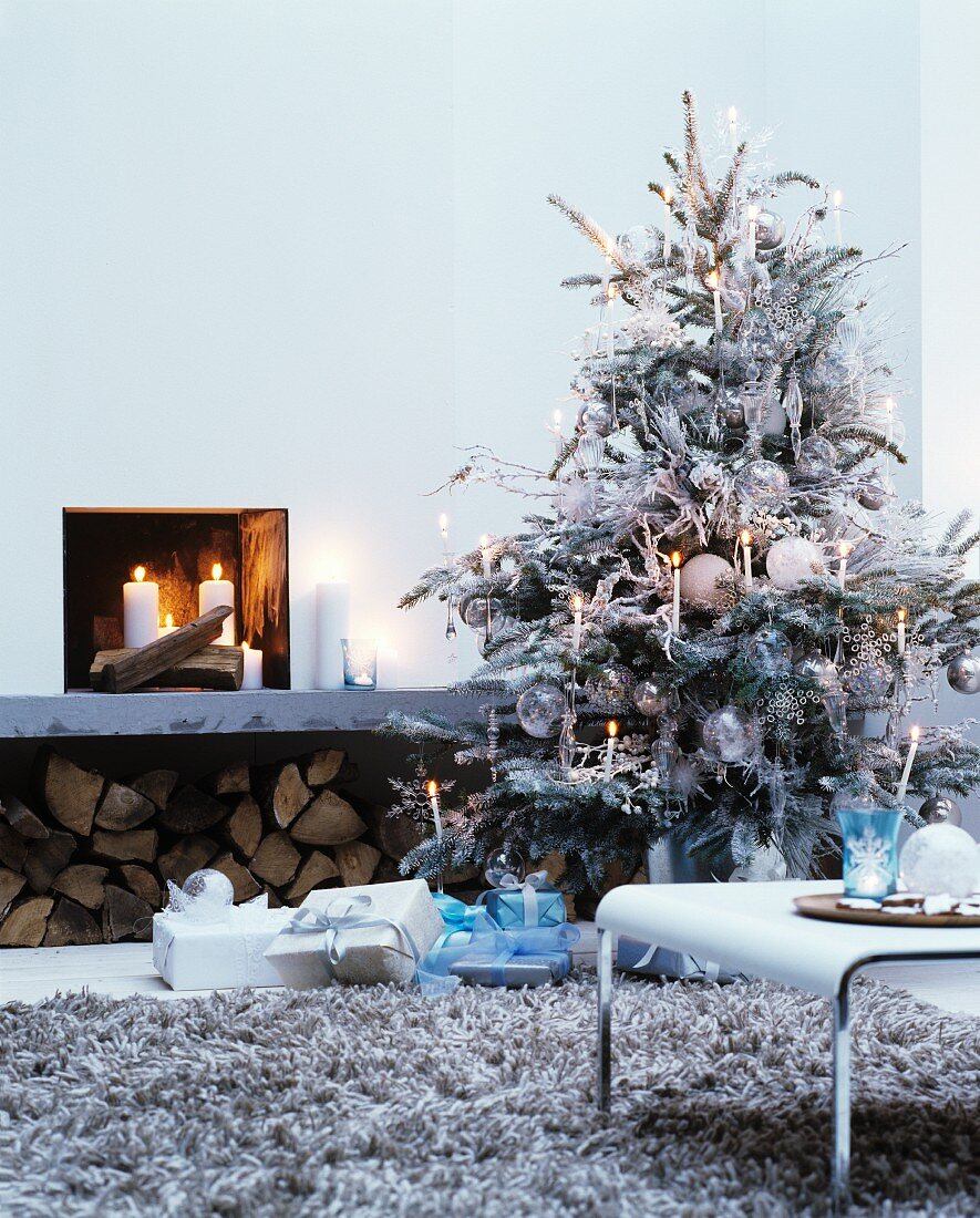 Weiß besprühter Christbaum mit weissen Kerzen und Glasschmuck dekoriert neben offenem Kamin